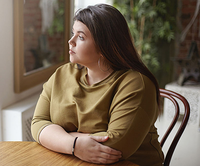 Una joven con sobrepeso sentada a una mesa y mirando por una ventana.