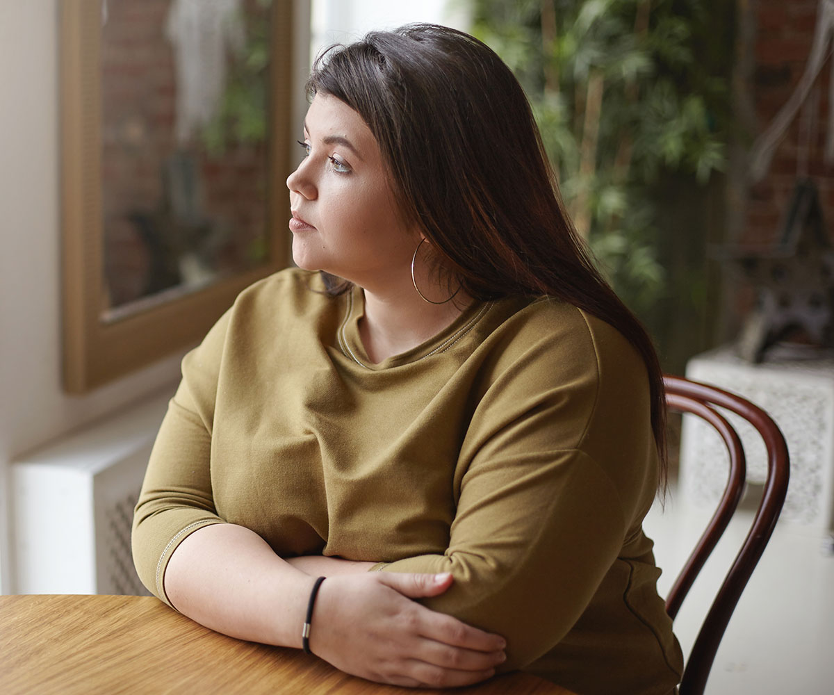 Una joven con sobrepeso sentada a una mesa y mirando por una ventana.