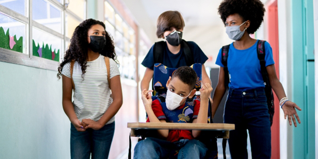 Un grupo de adolescentes camina por el pasillo de una escuela y llevan a un estudiante en silla de ruedas por el pasillo. Todos usan mascarillas.