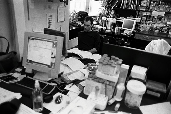 Fotografía en blanco y negro que muestra un escritorio cubierto de papeles, tubos de ensayo y una computadora en primer plano. Un hombre sentado frente a la pantalla de una computadora con libros, equipos de investigación científica y una estación de trabajo adicional detrás de él.