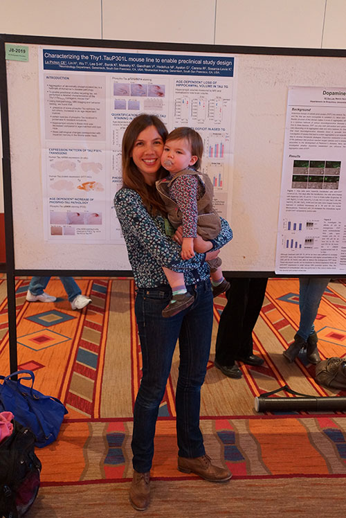 Una mujer con un niño pequeño en sus brazos delante de un cartel de investigación científica.