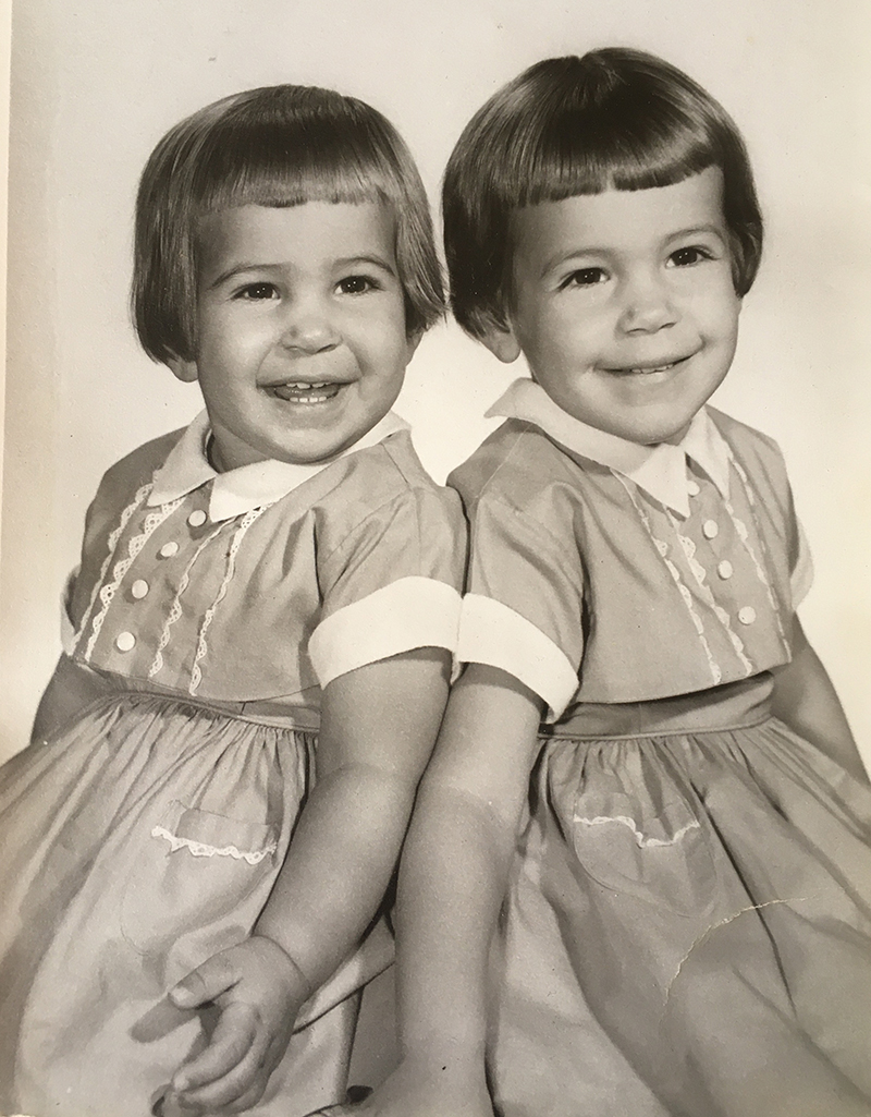 Dos chicas sonríen en una fotografía en tonos sepia. Llevan vestidos idénticos y tienen cortes de pelo idénticos.