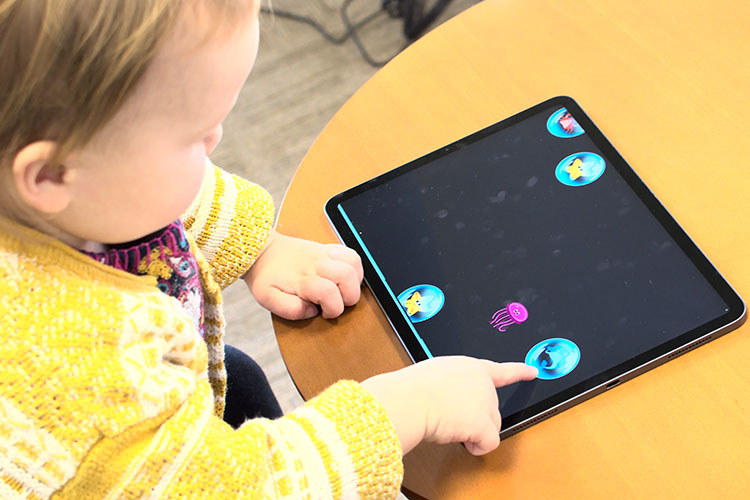 Una niña vestida con un suéter amarillo y sentada a una mesa utiliza la punta del dedo para hacer estallar una burbuja en una tableta.