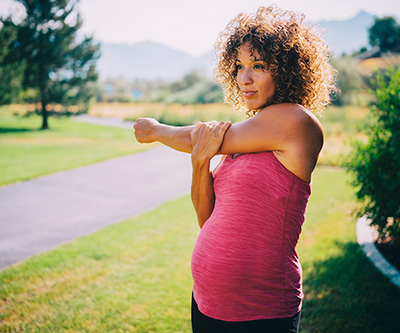 Una mujer embarazada con una camisa rosa estira su hombro en un parque con un sendero para correr.