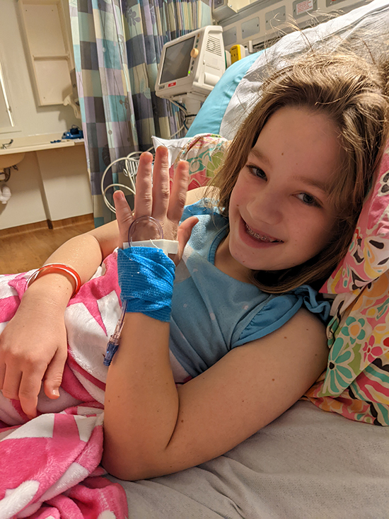 Una joven sonriente con una camisa azul yace en una cama de hospital. Ella levanta su mano, que está cubierta por un vendaje azul que sostiene una vía intravenosa en su lugar.