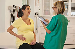 Una mujer embarazada está sentada en la mesa de examen en el consultorio del médico. Tiene su mano derecha sobre su estómago.  Una enfermera está de pie frente a ella y sosteniendo una computadora tipo tableta.