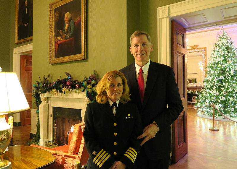 Una pareja sonríe para la cámara dentro de una habitación verde con chimenea. La Dra. Rouault lleva su uniforme del Servicio de Salud Pública. Hay un árbol de Navidad y decoración en la repisa.