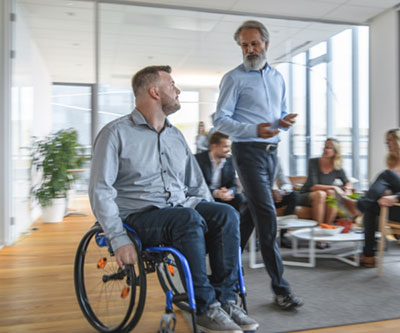 Un hombre en silla de ruedas conversa con otro hombre que está de pie. Ambos se desplazan por una habitación donde hay otros adultos sentados alrededor de una mesa de centro.