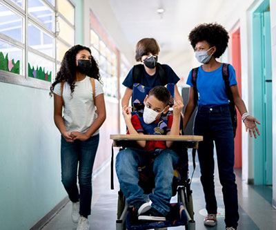 Un grupo de adolescentes camina por el pasillo de una escuela y llevan a un estudiante en silla de ruedas por el pasillo. Todos usan mascarillas.