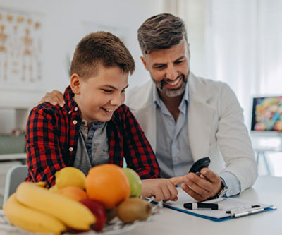 Un proveedor de atención médica verifica la glucosa en sangre de un adolescente mediante un pinchazo en el dedo con un monitor. Ambos sonríen y se ve un plato de frutas frescas sobre la mesa.