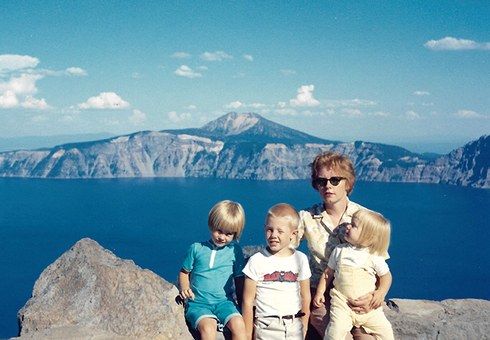 Tres niños de cabello rubio están sentados con su madre, que lleva gafas de sol, frente a Crater Lake.
