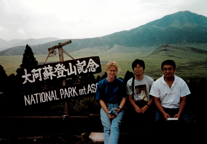 El grupo está sentado junto a un letrero de madera con letras blancas. La parte superior del letrero tiene escritura japonesa y la parte inferior dice: 'NATIONAL PARK mt ASO'; se puede ver la montaña en el fondo.
